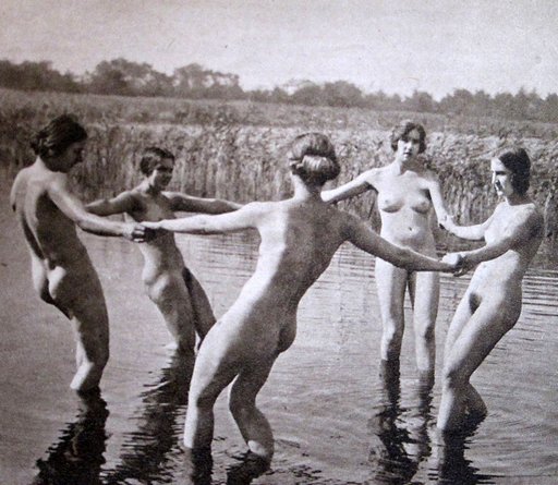 five naked white women holding hands and dancing in a circle in ankle-deep water