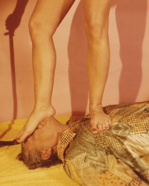 naked woman stepping on the face of an old man in silk pajamas