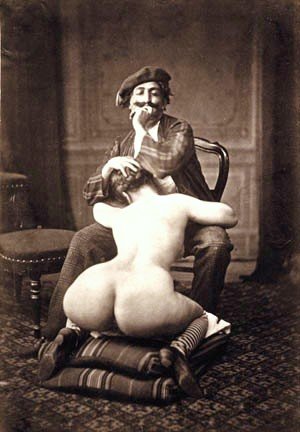 mustachioed man wearing a beret and sitting comfortably in a chair while getting a blowjob from a curvy woman who is kneeling before him on some cushions