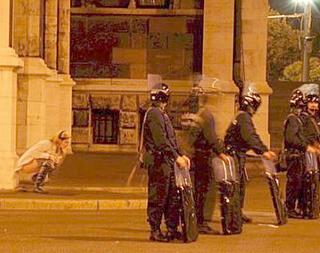 blonde woman squats to pee behind a line of riot police