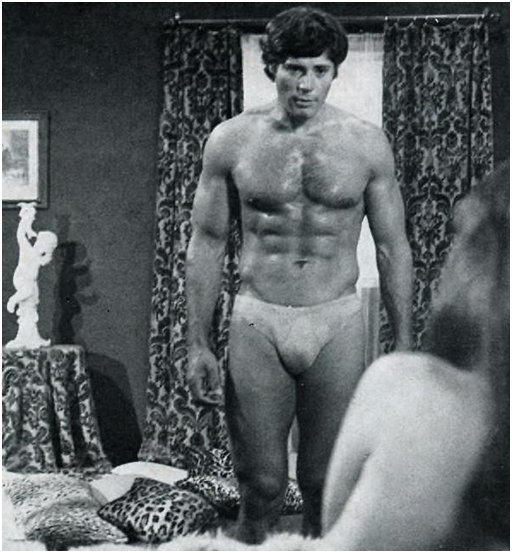 hunky man in speedo underwear looms over a topless model lounging in a bed