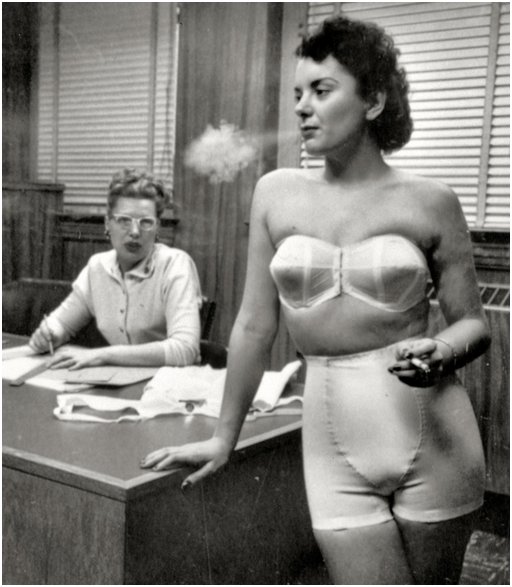 hard-looking brunette in vintage lingerie with cone bra and girdle type foundation undergarment, smoking a cigarette while an office lady secretary looks on