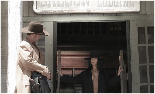woman in cowboy hat and trenchcoat with her breasts showing slightly comes out through some saloon doors to greet a lounging cowboy in a western scene shot in black and white
