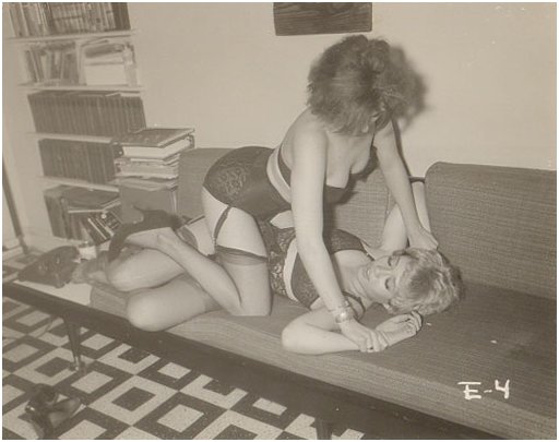 Two women in fancy lingerie are wrestling on a couch in this vintage photograph, watermarked E-4 in the style of Irving Klaw studios